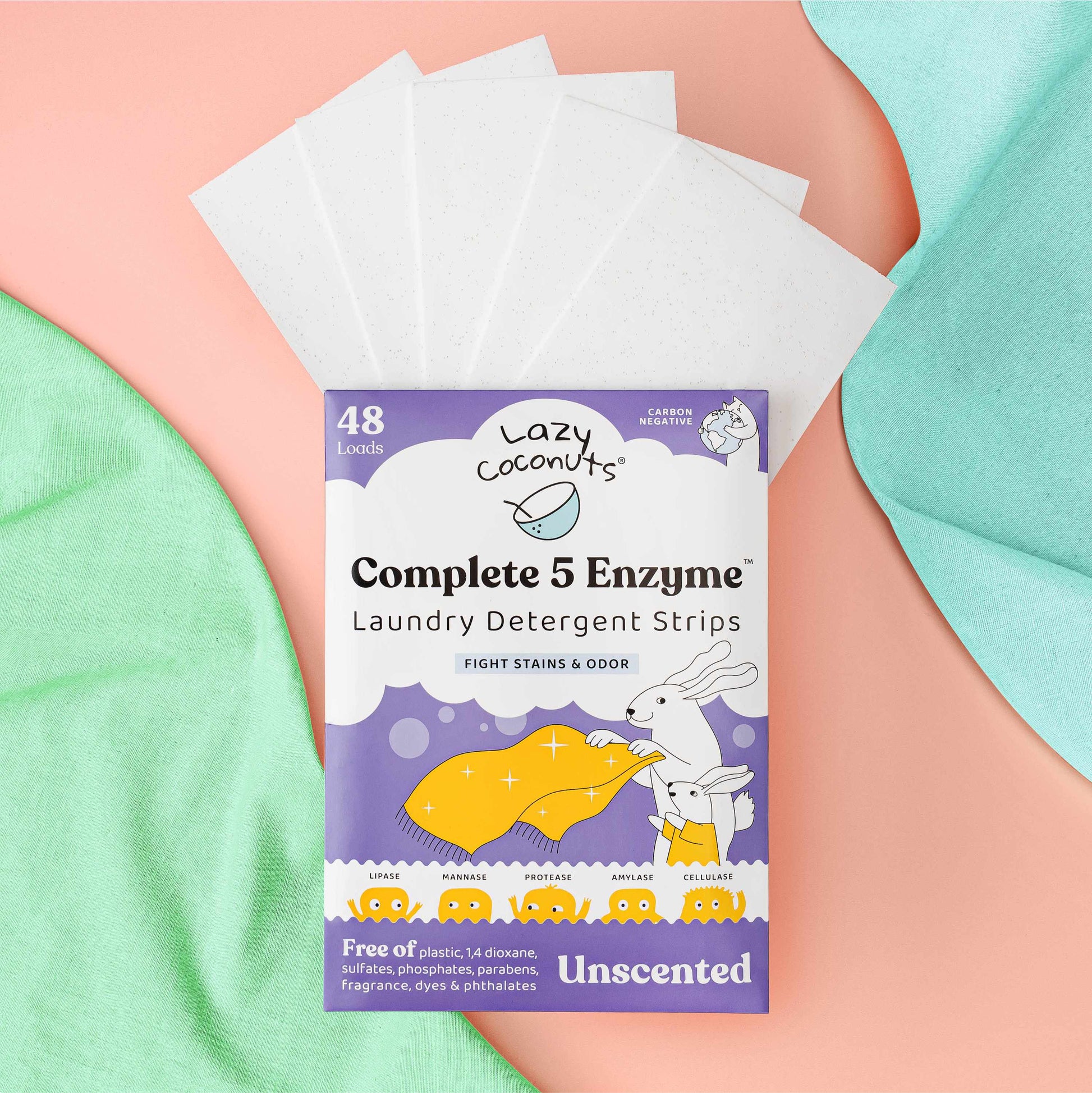Lazy Coconuts Complete 5 Enzyme Laundry Detergent Strips in a purple pouch with white detergent sheets fanned out behind it, styled on pastel pink and mint green fabric.