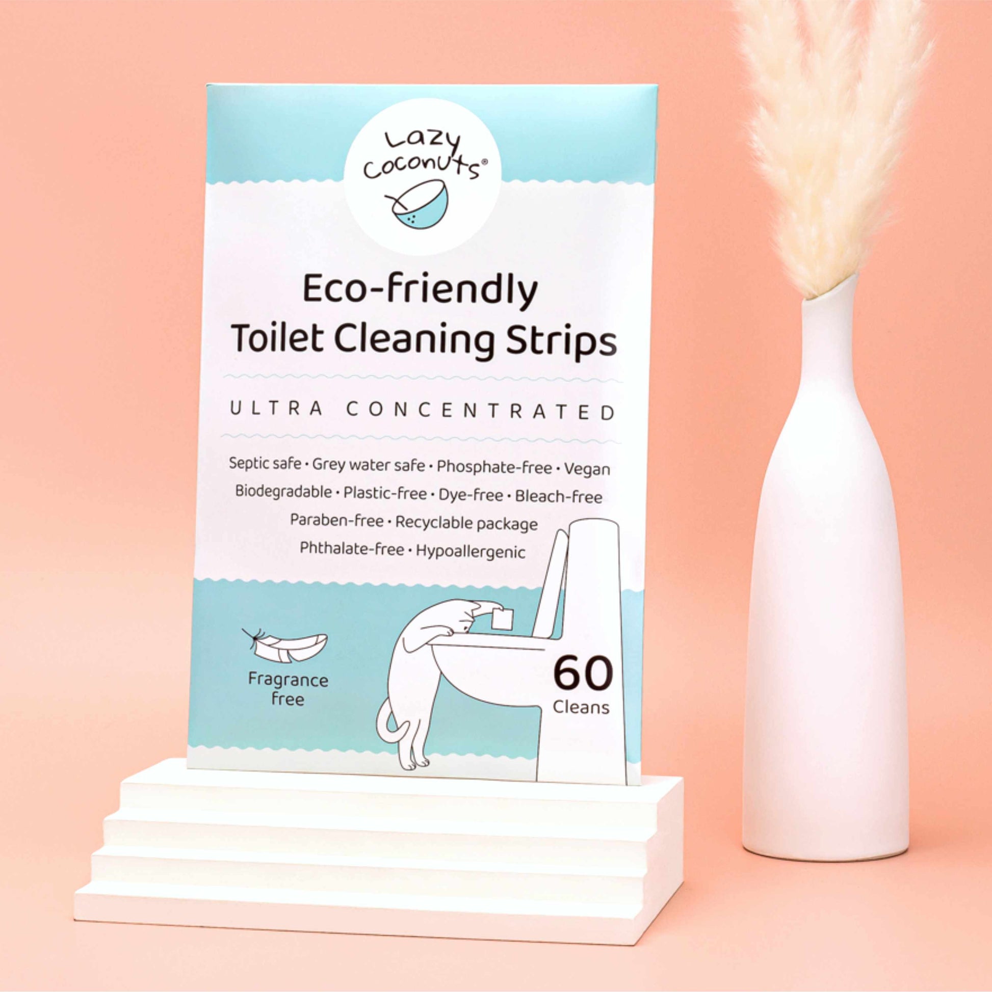 Lazy Coconuts Eco-Friendly Toilet Cleaning Strips box displayed on a white pedestal next to a white vase with dried pampas grass, set against a solid peach-pink background.
