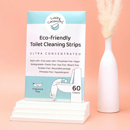 Lazy Coconuts Eco-Friendly Toilet Cleaning Strips box displayed on a white pedestal next to a white vase with dried pampas grass, set against a solid peach-pink background.