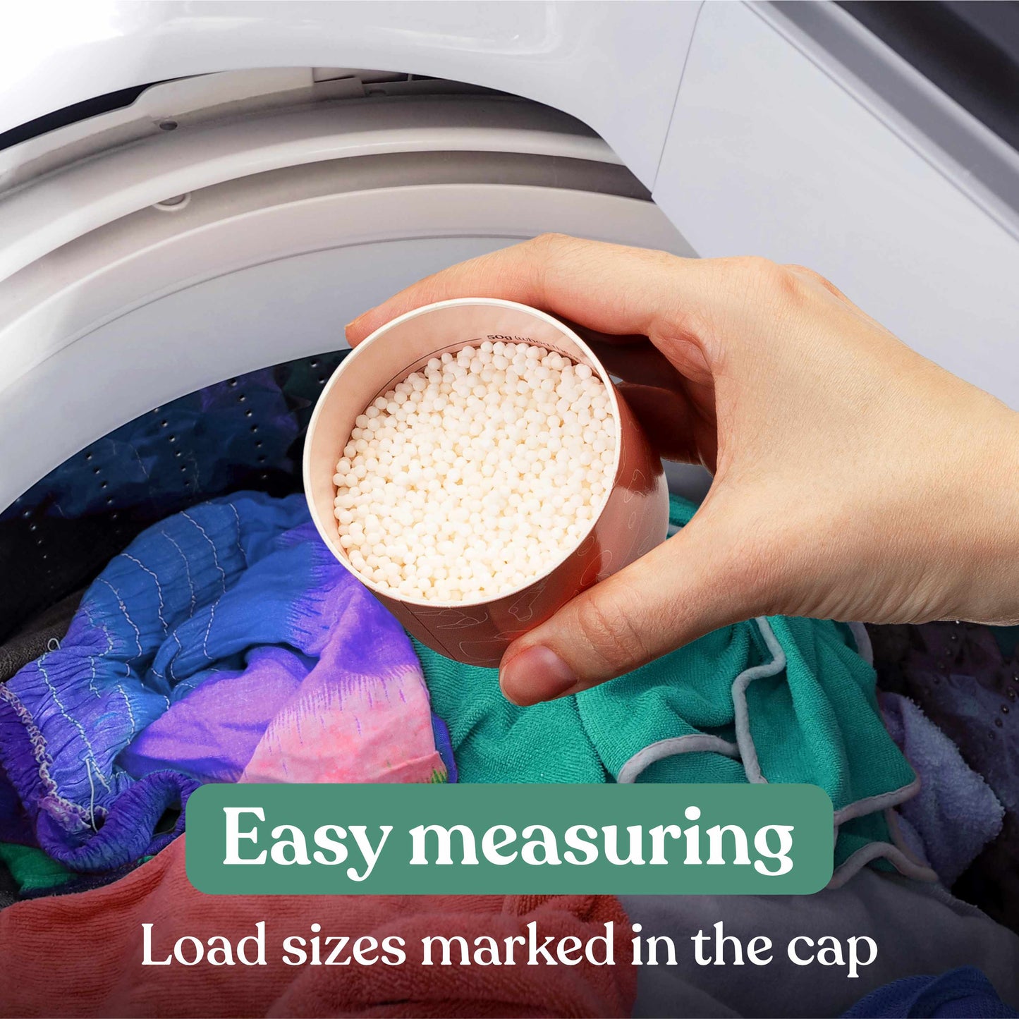 A hand holds a small cup filled with white laundry scent booster granules in front of an open washing machine door with colorful clothes inside. Text overlays read 'Easy measuring' and 'Load sizes marked in the cap'.