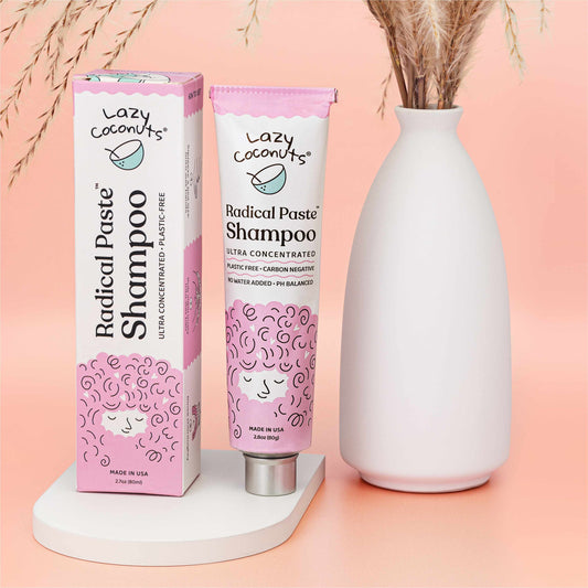 Lazy Coconuts Radical Paste Shampoo in a pink squeeze tube with its matching pink and white box, styled on a white platform next to a white ceramic vase with dried grass against a soft peach-pink background.