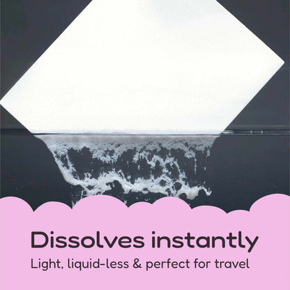 A square white detergent strip shown at an angle over a dark reflective surface, with foamy dissolution at the bottom and pink text reading 'Dissolves instantly' and 'Light, liquid-less & perfect for travel'.