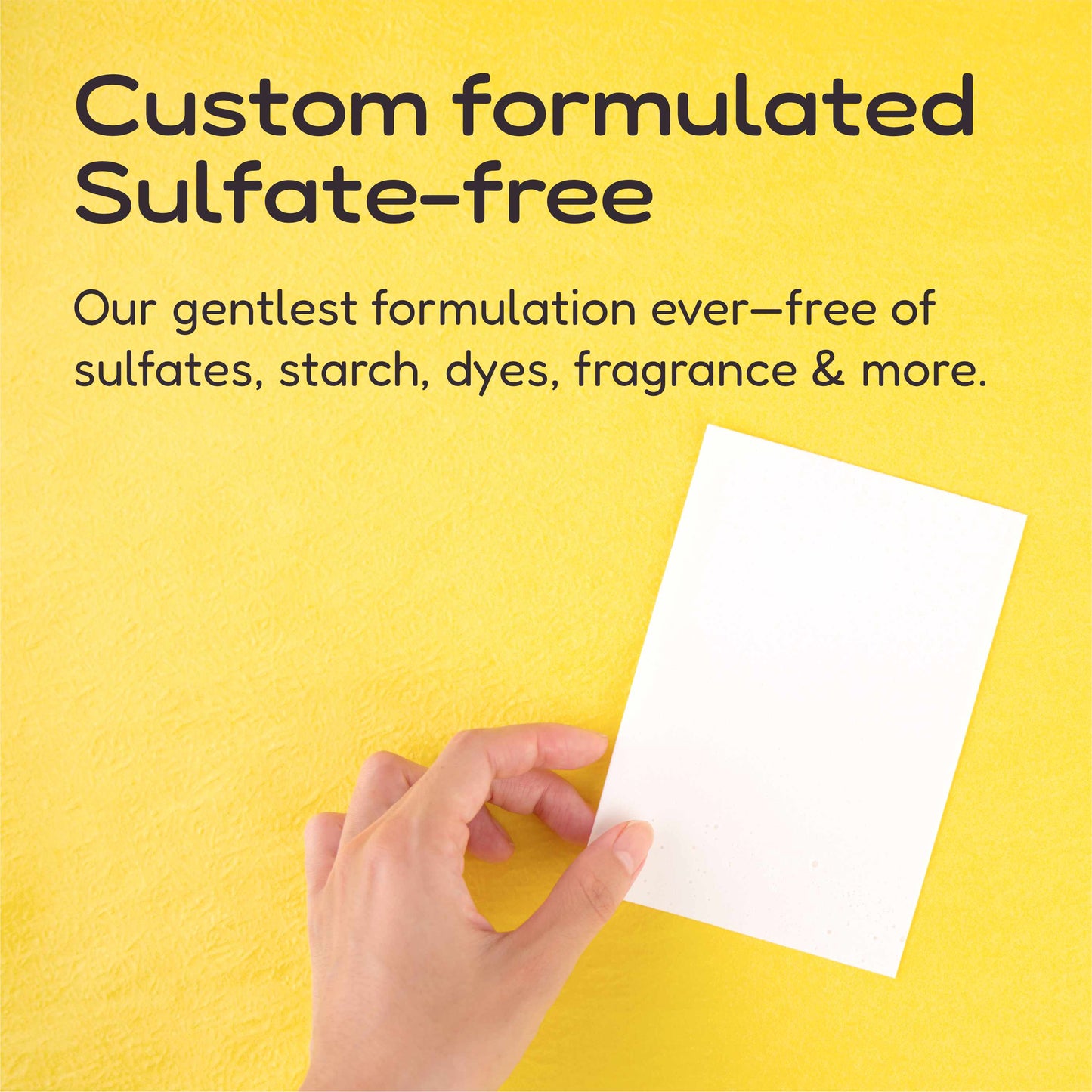 A hand holds a white laundry detergent sheet against a bright yellow background with black text reading 'Custom formulated Sulfate-free' and additional descriptive copy about being free of sulfates, starch, dyes, fragrance & more.